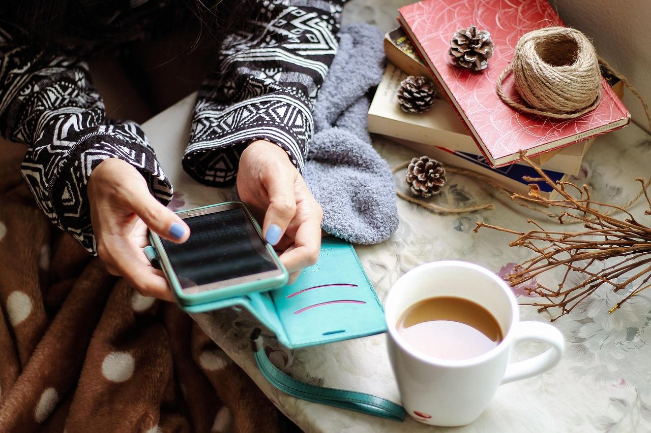 Consumer using smartphone with coffee and books nearby, representing enhanced contact information like updated emails, phone numbers, and mailing addresses for holiday campaigns.